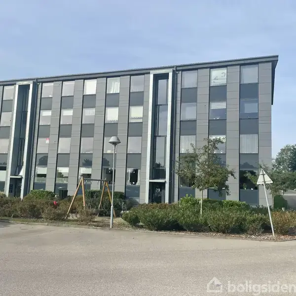 En fire-etagers kontorbygning står stille med glasvinduer i rækker. Foran er en parkeringsplads og grønne buske. Sollys belyser den åbne plads, mens enkelte træer omkranser området.