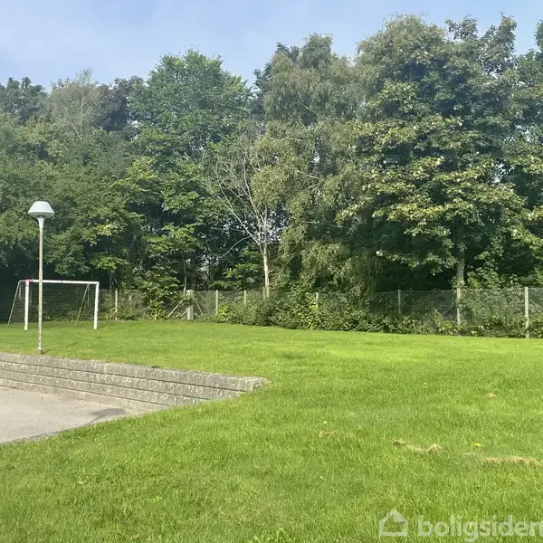 Fodboldmål står stille ved siden af en asfaltsti; omgivet af grønne græsplæner og tætte træer i baggrunden.