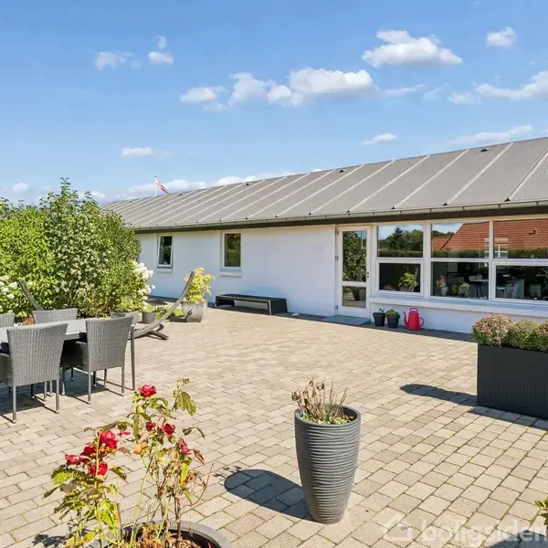 Terrasse med hvide fliser, der indeholder et sort spisebordssæt og blomsterkrukker. Området er placeret foran et hvidt hus med store vinduer under en blå himmel med spredte skyer.