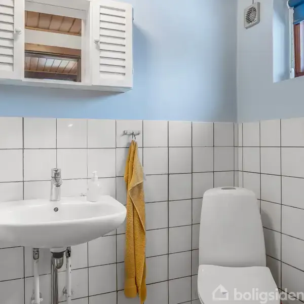 Håndvask med hane monteret på væggen; gul håndklæde hænger ved siden af. Toilet ved siden af, alle omgivet af hvide fliser på væggen og et lille vindue med blå rullegardin.