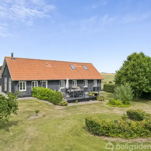 Et gråt hus med rødt tegltag står i en grøn have med buske og træer. Huset har en terrasse med havemøbler, og landskabet omkring er åbent og landligt.