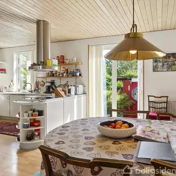 Et hyggeligt køkken med et rundt spisebord dækket af en dug og frugter i en skål. Køkkenet har hvide skabe, en emhætte og vinduer, der lukker lys ind. Der er også en udgang til en have.
