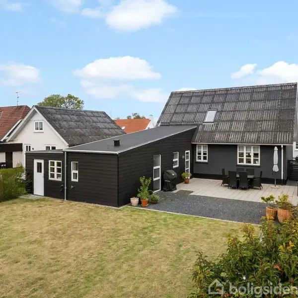 Sortmalet hus med gråt tag står i en baghave med en terrasse med havemøbler og planter i potter. Omgivet af græsplæne og naboernes huse i et villakvarter.