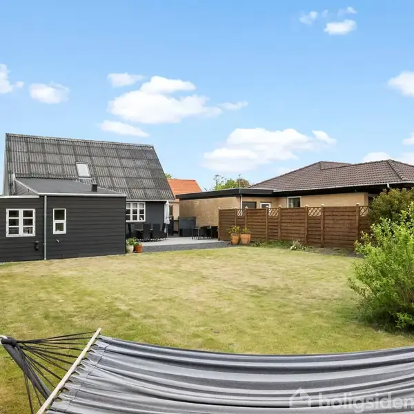 Hængekøje står stille i en velplejet græsplæne. I baggrunden ses et sort-hvidt hus og et ryddeligt træhegn under en blå himmel med få skyer.