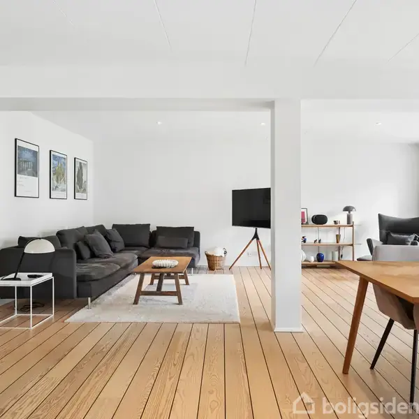 En stue med en sort sofa og sofabord placeret på et lyst tæppe. Til højre står et spisebord med stole. Væggene er hvide, og der hænger tre billeder.