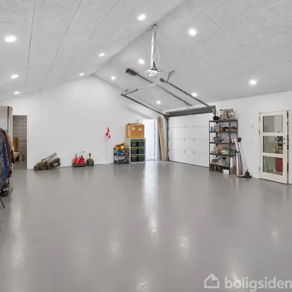 En rummelig garage med høj loftshøjde og gråt gulv. Hylder, værktøj og opbevaringskasser står langs væggene. Store garageporte og glasdøre er synlige i den lyse oplyste indretning.