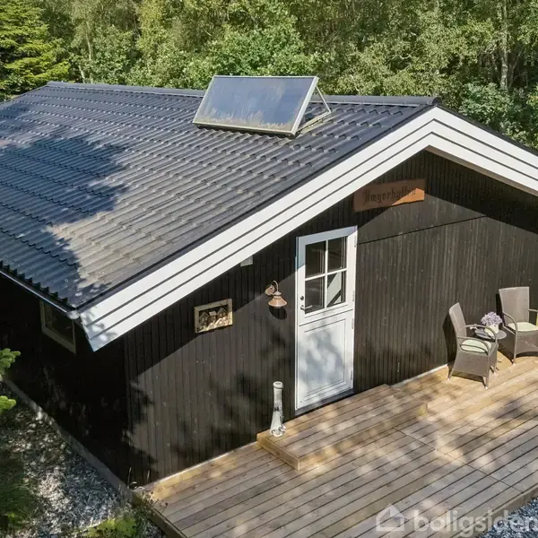 Sort træhus med tagvindue står på en træterrasse omgivet af skov. To stole og et bord står ved siden af hoveddøren. Skilt over døren siger "Ingeborgs hus".
