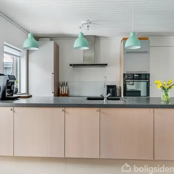 Køkkenet har en lang grå bordplade med indbygget vask, tre mintgrønne lamper hænger over bordet. Der er en elkedel, kaffemaskine, ovn og en vase med blomster til højre.