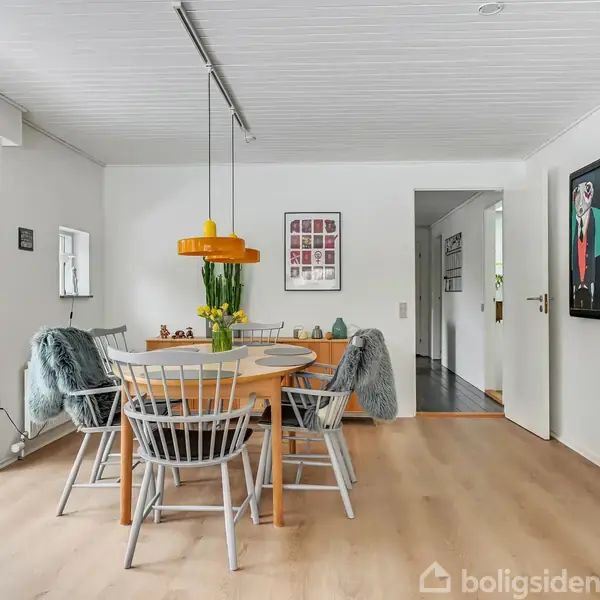 Rundt spisebord med træstole omringet af hvidmalede vægge. Over bordet hænger en gul lampe. Malerier på væggene og store glasdøre leder ud til en have.