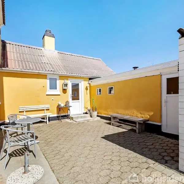 En gulmalet gård med bænke og bistrostole arrangeret på terrassefliser. Solskin lyser op mod huse med røde tegltage. En lille hvid træbygning står til højre.