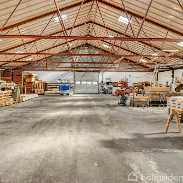 Lagerhal med træplanker og værktøj opbevaret på hylder og arbejdsborde, ligger under et højt, spidst tag. Store åbne døre i baggrunden giver adgang til dagslys.