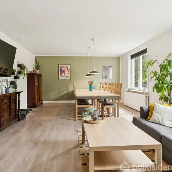 En stue med et gråt hjørnesofa og en træsofabord, placeret ved siden af et stort vindue. Et spisebord med flere stole står i baggrunden ved en grøn væg.