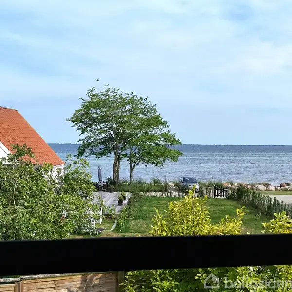 En hvid bygning med rødt tag står ved siden af en grøn have med træer. I baggrunden ses havet og en klar blå himmel.
