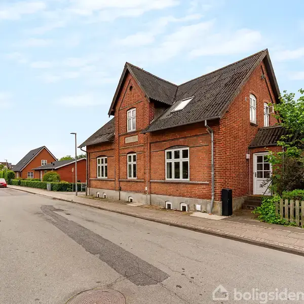 Rødt murstenshus står langs en stille gade med fortov, omgivet af buske og træer i en mindre by. To biler ses længere nede ad vejen.