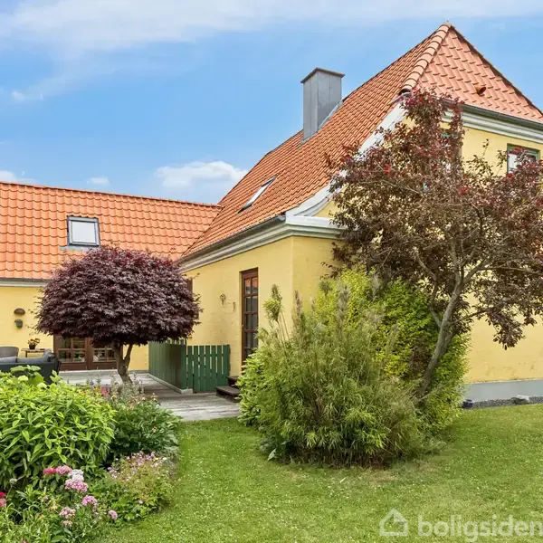 Gult hus med rødt tegltag står i en velplejet have med blomster og buske. I baggrunden viser himlen blå nuancer med få skyer.