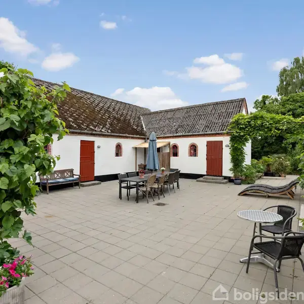 Gårdhave med flisebelægning, møbleret med bord, stole og en parasol. Omgivet af hvidmalede bygninger med røde døre og grøn beplantning, herunder blomster og buske.