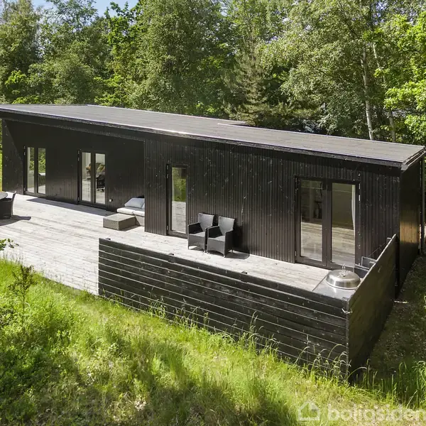 Sort træhus med store vinduer står på en rummelig terrasse med havemøbler, omgivet af frodig skov.
