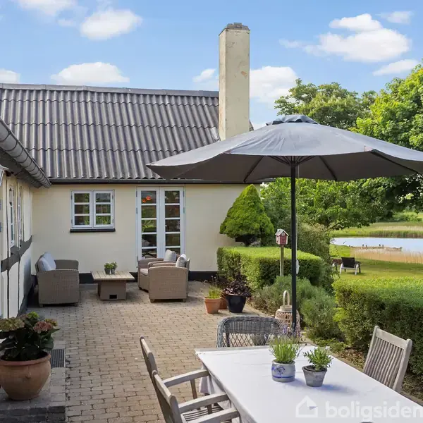 Hus med terrasse møbleret med borde og stole under parasol; i baggrunden ses en have med græsplæne, træer og udsigt til en sø i naturlige omgivelser.