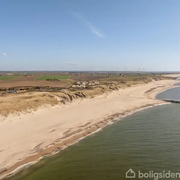 En lang, hvid sandstrand strækker sig langs kysten med klitter og grønne marker i baggrunden. Havet ligger til højre, med enkelte bølgebrydere strækkende ud i vandet.