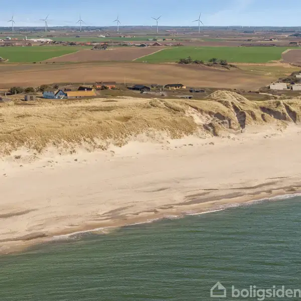 Strand med sandklitter ligger langs kystlinjen, hvor bølger møder land. I baggrunden ses spredt bebyggelse og flade marker med vindmøller i horisonten under en klar himmel.
