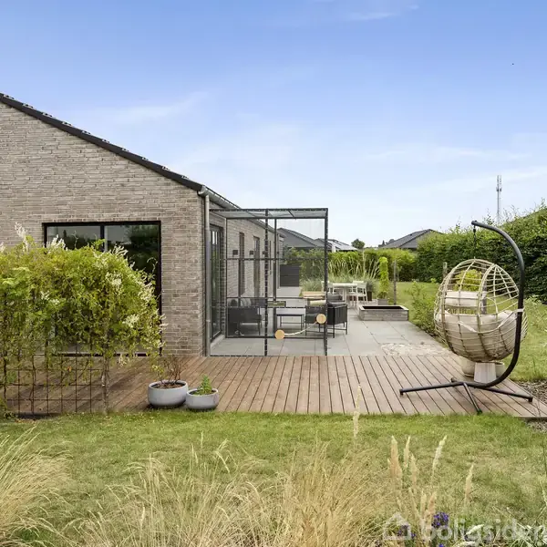 Et moderne hus med terrasse omgivet af frodige planter. Terrassen har et loungesæt og hængestol, mens en stenbygning danner baggrunden. Græs og buske omkranser området under en klar blå himmel.