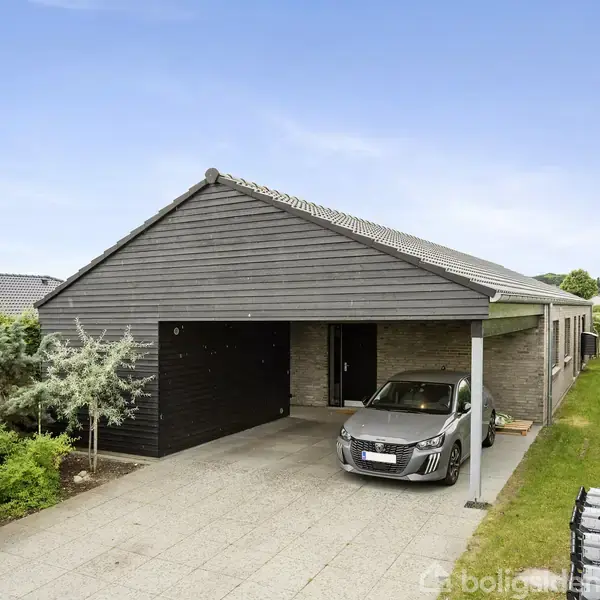 En moderne bil holder parkeret under en carport ved siden af et sort, træbeklædt hus. Husets forhave er anlagt med grønt græs og buske under en blå himmel.