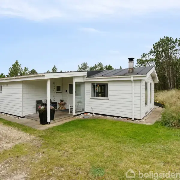 Et hvidt træhus med fladt tag står stille på en grøn græsplæne omgivet af træer. Huset har en overdækket terrasse og en grusvej foran.