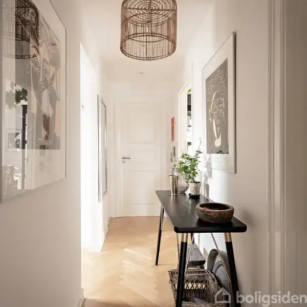 En smal gang med en sort bord, dekoreret med planter og skåle, hænger kunst på de hvide vægge. Gangen er lyst op af naturligt lys fra baggrundens dør.