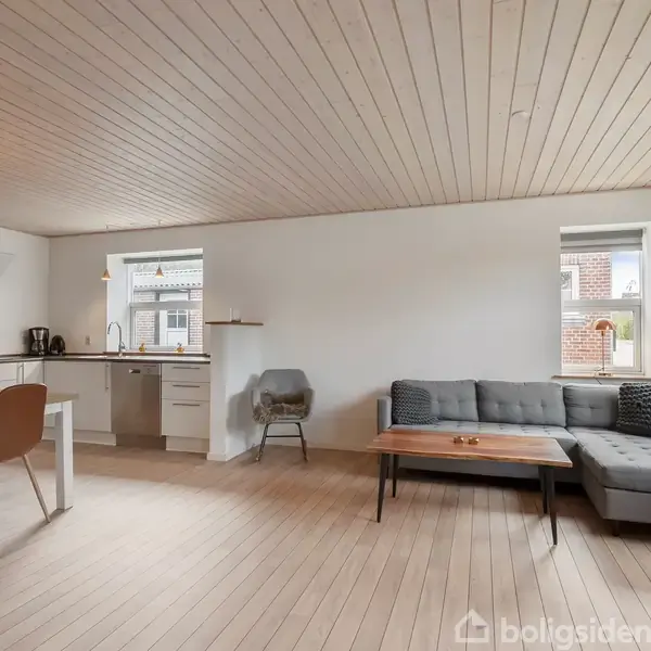 Stue med grå sofa og træbord står ved vindue, åben forbindelse til køkken med spiseplads, lyse trægulve og hvidmalede vægge.