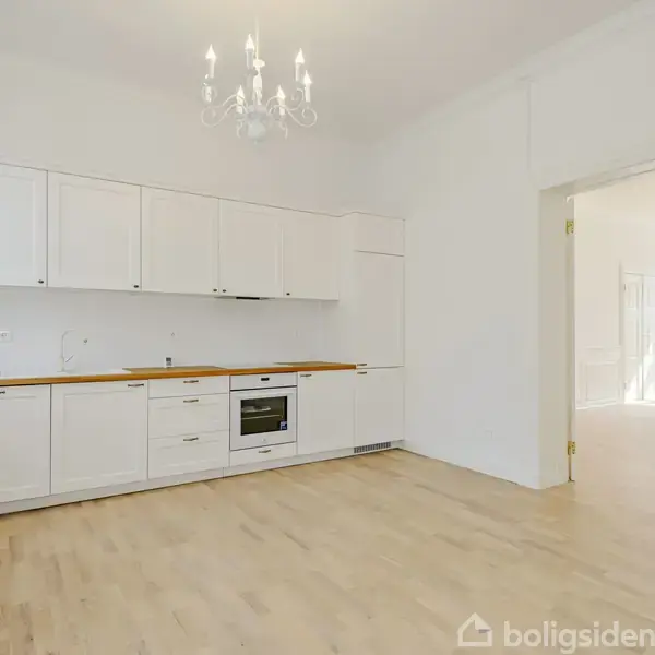 Et hvidt køkken med integreret ovn og træbordplade står i et rum med lyse trægulve og en lysekrone, der hænger fra loftet. Der er adgang til et tilstødende, oplyst værelse.