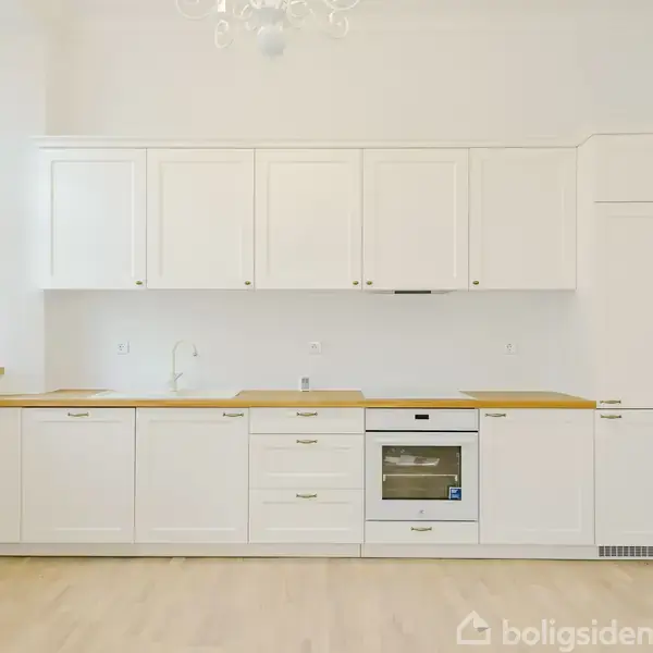 Et moderne køkken med hvide skabe og træbordplader står i et lyst rum. En ovn er indbygget i midten af underskabene. Store vinduer til venstre giver naturligt lys.
