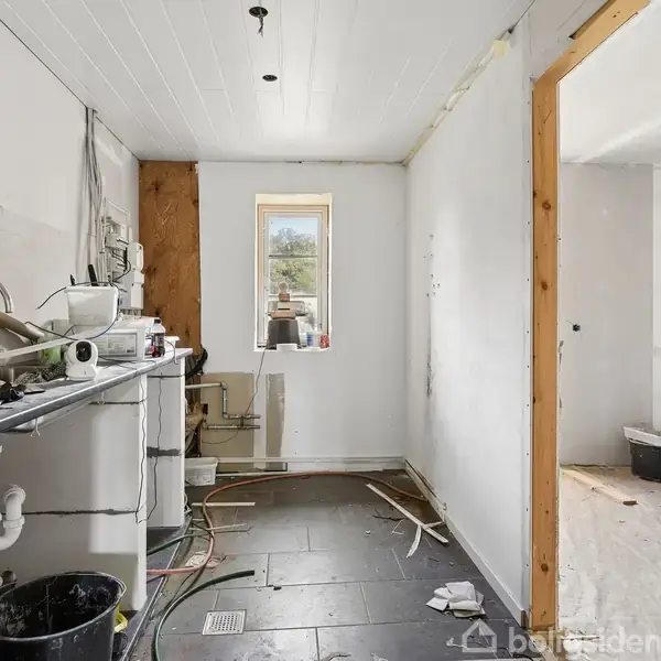 Et køkken under renovation med værktøj og byggematerialer på bordpladen. Der er fliser på gulvet og en enkeltstående vindue. Et tilstødende rum er synligt gennem en døråbning.