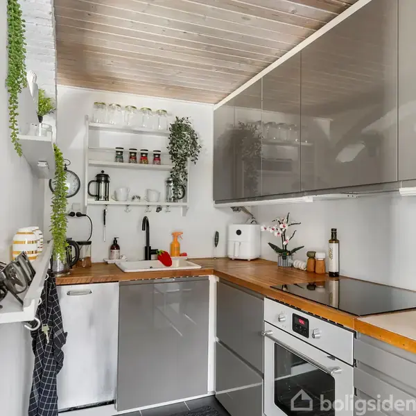 Køkken med grå skabe og træbordplade. Glas og krydderiglas står på hylder, mens planter hænger på væggene. En ovn og kogeplade er indbygget. Luce belysning ovenover.