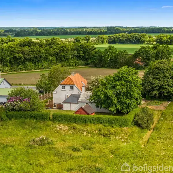 Et hvidt hus med rødt tag står omgivet af frodige træer og grønne marker, der strækker sig mod en skov i baggrunden.