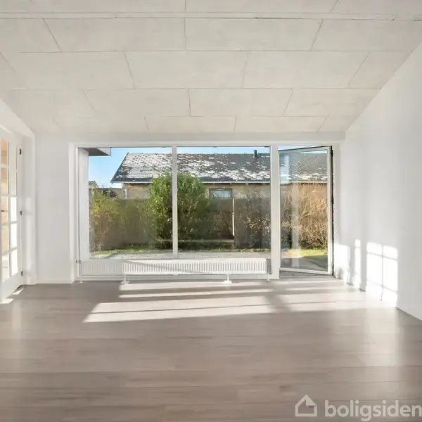 Stor tom stue med lyse trægulve, store vinduer med udsigt til en have og hvidmalede vægge. Sollys strømmer ind og fylder rummet.