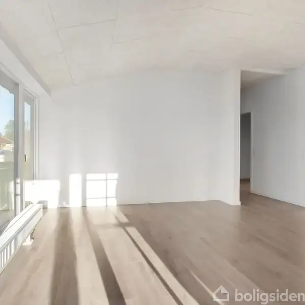 Stue med træparketgulv, hvor sollys strømmer ind gennem store vinduer til venstre. I baggrunden er der et hvidt køkken og åbent rum.