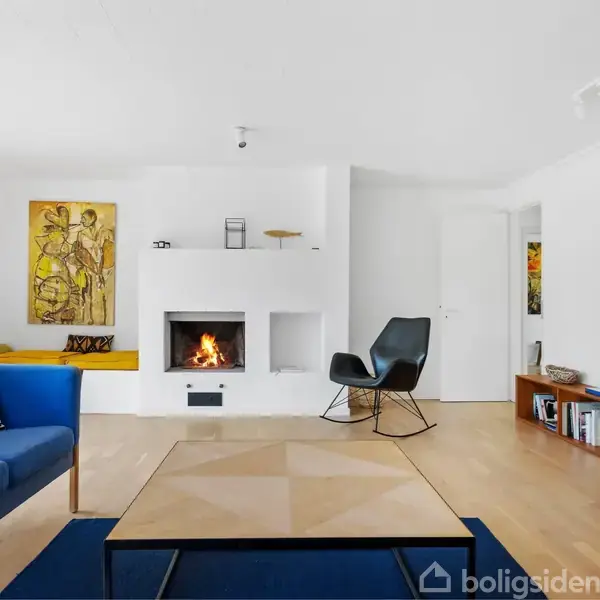 Blå sofa og sort stol står i en stue med hvid pejs, hvor ilden brænder. Væggene er dekoreret med farverige malerier, og der er en bogreol i hjørnet.