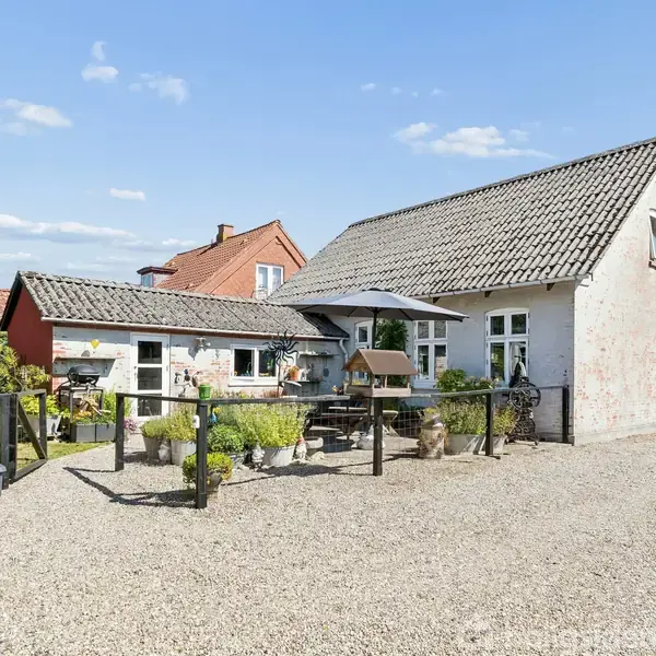 Et hvidt stuehus med skråtag står på en grusbelagt gårdsplads. Foran huset er der en parasol, planter og en træhavebænk. Omkringliggende grønne buske og lidt blå himmel.