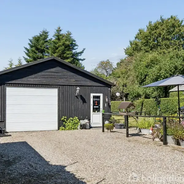 En sort bygning med en hvid garageport og dør er centreret. Foran er der en grusbelagt indkørsel med planter. Omkring er grøn beplantning og en solrig gårdhave med en parasol.