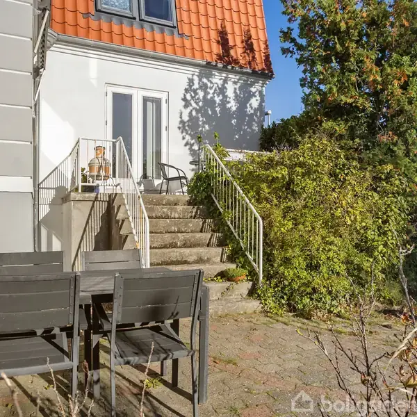 Hus med hvid facade og rødt tegltag, trappe fører op til altan med stoler, ved siden af terrasse med sort spisebordssæt, omgivet af grønne buske og træer i sollys.