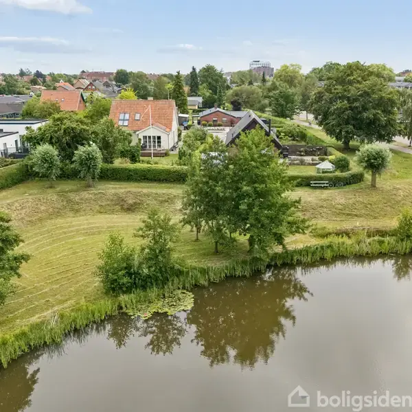En sø er omgivet af grønne træer og velplejede græsplæner. I baggrunden ses huse med røde tage og en lille by i horisonten under en blå himmel.