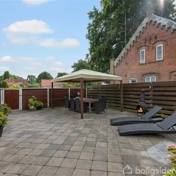 Terrasse med flisebelægning; to solsenge og et spisebord under en parasol. Grænset af et plankeværk og en murstensbygning med dekoreret gavl. Planter i krukker står langs væggen.