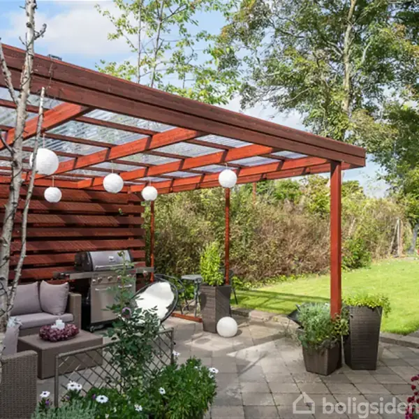 Overdækket terrasse med sofa og grill, dekoreret med hængende lanterner. Terrassen er omkranset af en flisebelagt have med blomsterkrukker og græsplæne. I baggrunden ses buske og træer.