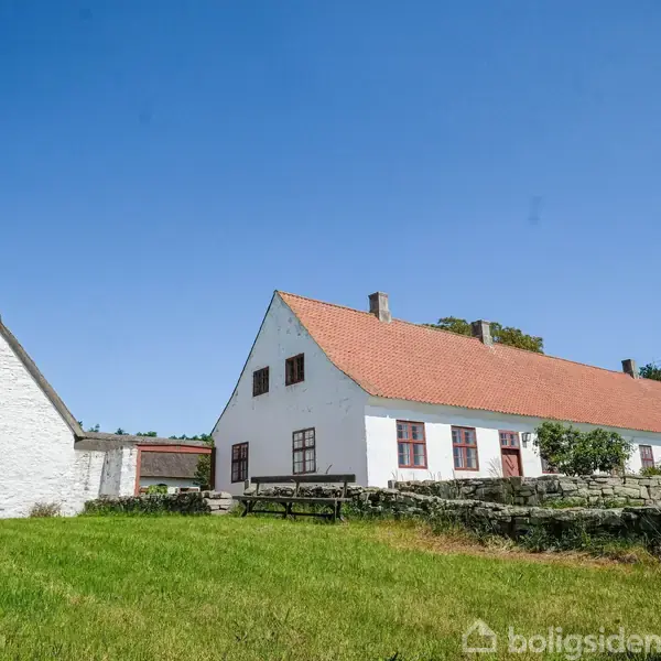 Hvid bygning med rødt tegltag står stille i en grøn mark omkranset af en stenmuret have under en klar blå himmel.