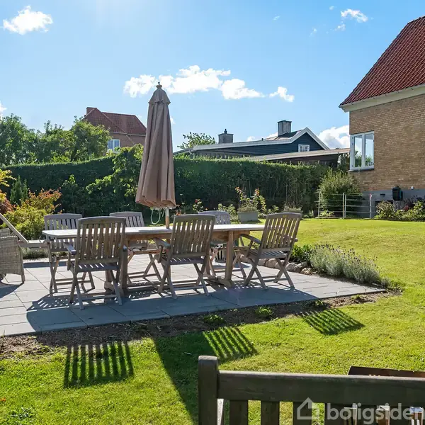 Et udendørs spisebordssæt står på en flisebelagt terrasse i en solrig have ved siden af et rødt murstenshus med en trappe. Tætklippet græsplæne omgiver området.