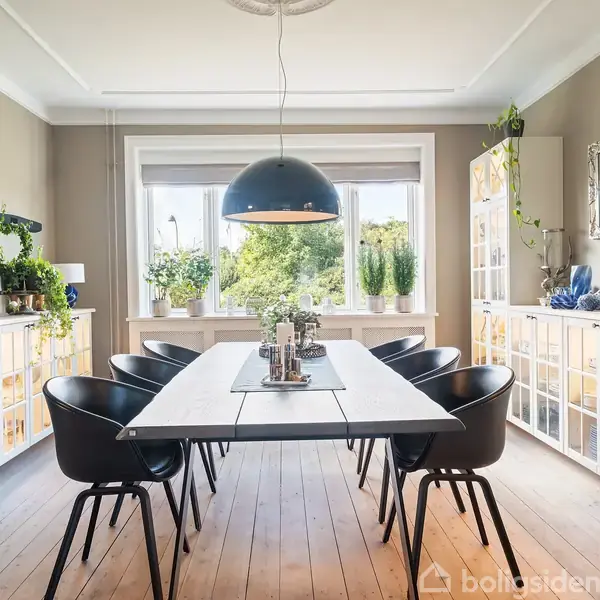 Et rektangulært spisebord står i midten af et lyst rum med sorte stole omkring. En stor lampe hænger over bordet. Væggene har hylder med planter og dekorationer.