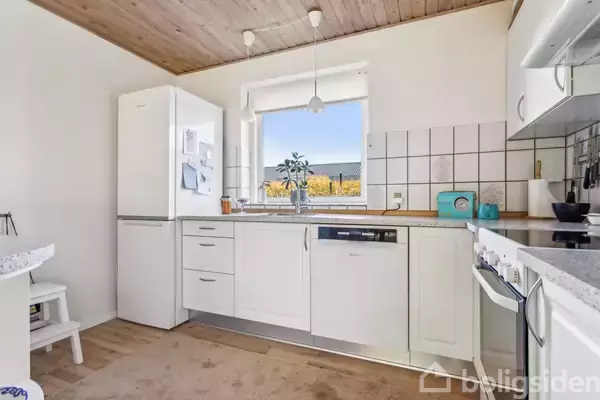 Køkken med hvide skabe inkluderer køleskab, opvaskemaskine og komfur. Et vindue med udsigt til haven bagved. En plante og opbevaringskrukker står på køkkenbordet. Spots i loftet.