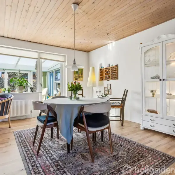 En spisestue med et rundt bord dækket med dug omgivet af stole. En hvid vitrine viser porcelæn. Rummet er lyst med trægulv og loft, set gennem store vinduer med udsigt til en terrasse.