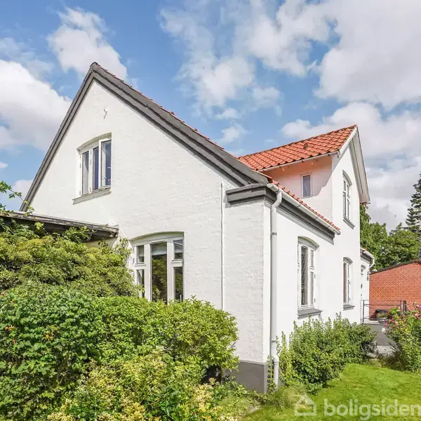 Hvidt hus med rødt tag ved siden af frodig have. Det står i et beboelsesområde med andre huse og træer i baggrunden under en blå himmel med skyer.