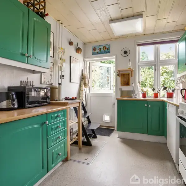 Et køkken med grønne skabe og træbordplader. Køkkenapparater, som en brødrister og mikroovn, står på bordet. Store vinduer og en glasdør giver udsigt til en have.